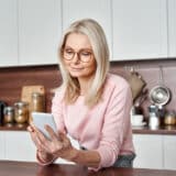 Mature blonde woman with glasses standing in her kitchen looking at her cellphone.