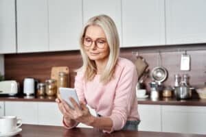 Mature blonde woman with glasses standing in her kitchen looking at her cellphone.