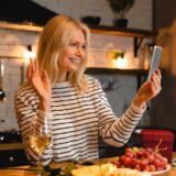 Blonde woman in a upscale kitchen smiling and waving into her phone.